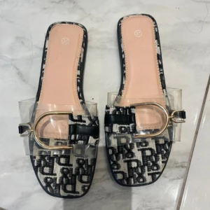 Svartvita sandaler med transparent rem - Snygga sandaler med fyrkantig tå, svartvitt mönster och transparent rem över foten. Dekorativt guldfärgat spänne framtill. Sulan är svart och ovandelen har ett grafiskt mönster. Perfekta för dig som vill sticka ut med en trendig look.