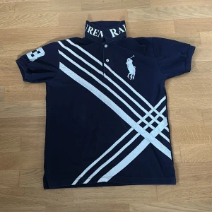 Mörkblå Polo Ralph Lauren piké  - Snygg mörkblå piké  från Polo Ralph Lauren med vita diagonala ränder och stor logga på bröstet. Kragen har tryckt text och ärmarna är korta med en broderad siffra på ena ärmen. Klassisk piké-look i bomull, perfekt för en sportig stil. Sitter smått  i storlek 