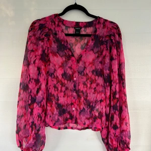 Blommig rosa blus - Snygg blommig blus från Lindex i transparent material. Färgerna går i rosa, lila och rött med v-ringning, knappar framtill och långa puffärmar. Perfekt för dig som vill sticka ut med färg och mönster.🤩💕