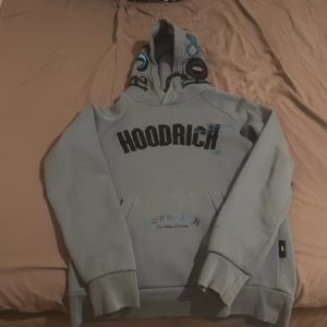 Ljusblå  hoodie från Hoodrich XS - Säljer en ljusblå hoodie från Hoodrich i storlek XS. Tröjan har stor svart logga framtill, tryck på fickan och text på ryggen. Huva med tryck och klassisk känguruficka. Perfekt för dig som gillar streetwear och vill sticka ut.
