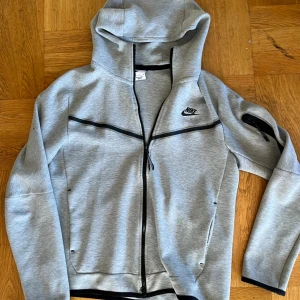Grå Nike tech fleece hoodie M - Snygg grå hoodie från Nike i storlek M med hel dragkedja och svarta detaljer. Tröjan har huva, långa ärmar och praktiska fickor. 