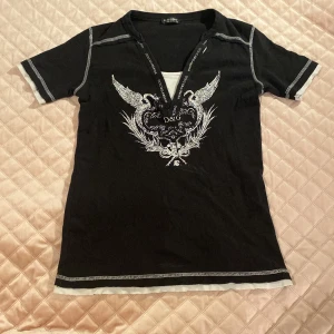 Svart D&G t-shirt med v-ringning - Svart t-shirt från Dolce & Gabbana med vit print framtill och cool v-ringad hals med logoband. T-shirten har vita kontrastsömmar och detaljer vid ärmslut och nederkant. Snyggt tryck med vingar och D&G-logo på bröstet. Bra kvalitet och har aldrig använt den.