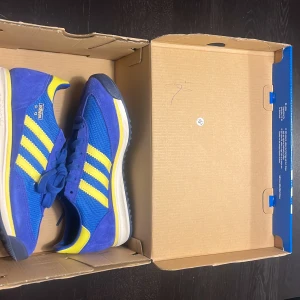Adidas SL 72 RS J blå/gula sneakers - Säljer ett par Adidas SL 72 RS J sneakers i blått och gult med klassiska tre ränder på sidan. Ovandelen är i mesh och mocka. Säljer på grund av fel storlek.Orgnal pris 900kr säljer för 799 