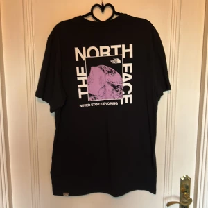 Svart t-shirt The North Face - Svart t-shirt från The North Face med stor vit och lila grafisk print på baksidan och liten logga på bröstet. Klassisk rund hals och korta ärmar. Skön passform och tillverkad i mjuk bomull. Perfekt för dig som gillar streetwear och snygga tryck.