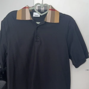 Svart Burberry t-shirt med rutig krage - Svart t-shirt från Burberry med klassisk rutig krage i beige, rött och vitt. Modellen har korta ärmar och knäppning framtill med tre knappar. Perfekt för dig som vill ha en stilren look med en touch av lyx.