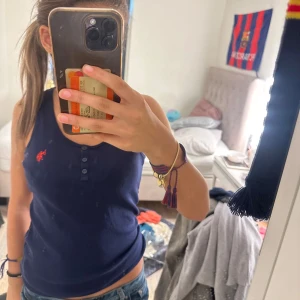 Mörkblå linnetopp med knappar - Säljer en mörkblå linnetopp med tre knappar framtill och en liten röd broderad detalj på bröstet. Toppen är ärmlös och har en klassisk, enkel design som passar perfekt till jeans eller shorts.