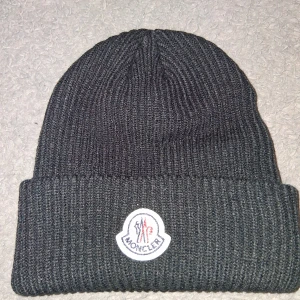 Svart ribbstickad Moncler mössa - Svart ribbstickad mössa från Moncler med klassisk uppvikt kant och broderad logotyp framtill. Tillverkad i mjukt material som håller dig varm under kyliga dagar. Perfekt accessoar för att lyfta din vinterstil.