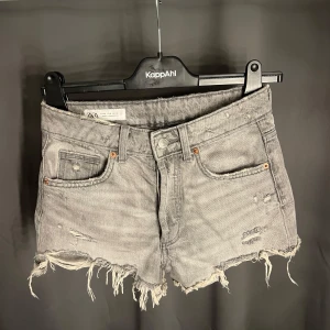 Grå jeansshorts med fransig kant från Zara - Grå jeansshorts från Zara med slitna detaljer och fransig rå kant. Klassisk femficksmodell, hög midja och knäppning med knappar. Perfekta för en avslappnad och trendig look i sommar.