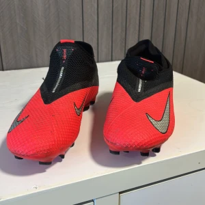 Nike Phantom Vision röda fotbollsskor - Säljer ett par Nike Phantom Vision fotbollsskor i rött och svart med silverfärgade swoosh-loggor. Skorna har en strumpliknande passform utan synlig snörning och är tillverkade i syntetmaterial med texturerad yta för bättre bollkontroll. Perfekta för dig som vill sticka ut på planen.