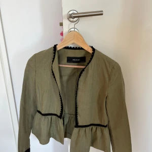 Grön omlottjacka från Zara Basic - Snygg grön omlottjacka från Zara Basic med svarta dekorativa detaljer längs kanterna och midjan. Jackan har volang nedtill och är tillverkad i linne, vilket ger en skön och luftig känsla. Långärmad och med en liten krok för stängning framtill.