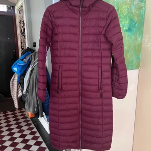 Vinröd lång dunjacka från Strova XS - Säljer en vinröd lång dunjacka från Strova i storlek XS. Jackan har huva, två sidofickor med dragkedja och quiltad design. Perfekt för kalla dagar med sin lätta och värmande känsla. Stängs med hel dragkedja framtill och har långärmade ärmar.