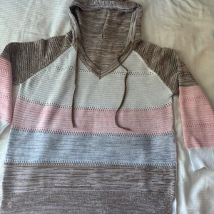 Randig hoodie i pastellfärger - Superfin stickad hoodie med breda ränder i beige, grått, vitt och ljusrosa. Tröjan har huva med snörning, v-ringning och långärmade ärmar. Perfekt för dig som gillar en chill och mysig stil med pastellvibbar.