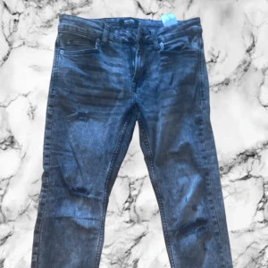 Gråa slitna jeans med raka ben - Snygga mörkblå jeans med slitningar på knäna och klassisk femficksdesign. Jeansen har raka ben och normal passform, tillverkade i mjukt denimtyg. Perfekta för en avslappnad och trendig look.