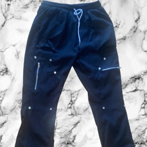 Svarta täckbyxor med dragkedjor - Svarta täckbyxor med flera silvriga dragkedjor och tryckknappar. Byxorna har resår och snörning i midjan för skön passform. Materialet är tåligt och passar perfekt för kalla dagar. Snyggt loose fit och praktiska fickor på benen.