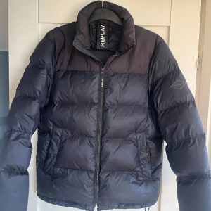 Varm och go svart pufferjacka från Replay - Säljer en svart pufferjacka från Replay med hög krage och dragkedja framtill. Jackan har två stora fickor på sidorna och är tillverkad i syntetmaterial. Perfekt för kalla dagar och har en clean, stilren look.