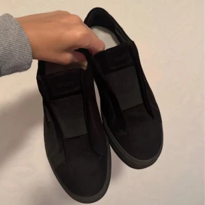 Svarta sneakers från Axel Arigato - Säljer ett par svarta sneakers från Axel Arigato i mocka med elastiskt band över vristen istället för snörning. Skorna har en stilren och minimalistisk design med rund tå och platt sula. Perfekta för dig som gillar en clean look och vill ha något som funkar till allt.