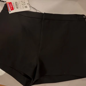 Svarta shorts med detaljer på sidorna  - Stilrena svarta shorts från pull and bear med snygg bältesdetalj i midjan och diskret bakficka. Klassisk rak modell som passar perfekt till en trendig look. Enkla att matcha med både t-shirt och skjorta.