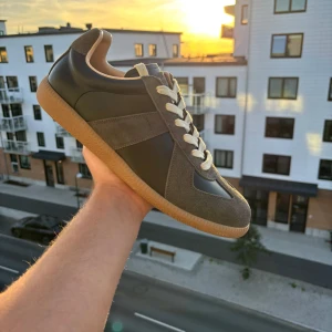 Maison Margiela - Snygga Maison Margiela sneakers med grå mockadetaljer och svarta partier i skinn. Skorna har vita snören och en klassisk gummisula i beige. Perfekt för dig som gillar stilrena och tidlösa sneakers med en retrovibe.