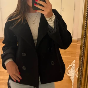 Svart kavaj, design by si - Stilren svart skepparkavaj från design by si med dubbelknäppning och bred krage. Jackan har stora fickor framtill, dekorativa knappar vid ärmslut och är tillverkad i ett mjukt ullblandat tyg. Perfekt för dig som gillar klassisk och clean stil.