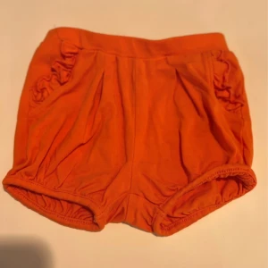 Orange shorts från Name It med volang - Snygga orange shorts från Name It i mjuk ekologisk bomull. Modellen är pösig med resår i midja och benslut samt volangdetaljer vid fickorna. Perfekta för varma dagar och superbekväma för de minsta.