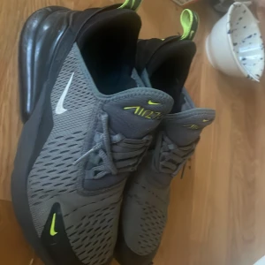Nike Air Max 270 grå/svart sneakers - Nike Air Max 270 sneakers i grått med svarta detaljer och neongula accenter. Skorna har en stor synlig Air Max-enhet i hälen, mesh-överdel för bra andningsförmåga och klassisk snörning. Perfekta för dig som gillar sportig och modern stil.