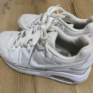 Vita Nike sneakers med snörning - Säljer ett par helvita Nike sneakers med klassisk design och synlig air-sula. Skorna har snörning, rund tå och är tillverkade i en mix av syntet och textil. Perfekta för dig som gillar en clean och sportig look.