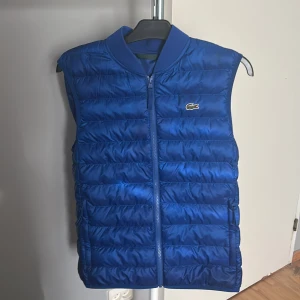 Blå dunväst från Lacoste - Snygg blå dunväst från Lacoste med klassisk krokodillogga på bröstet. Andvänd 2 gånger då den inte passar mig. Storlek M. Finns ej att köpa längre ny.