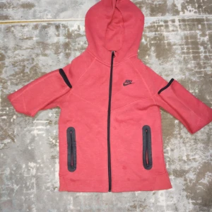 Röd hoodie från Nike med dragkedja - Nike hoodie i rött med svarta detaljer och hel dragkedja framtill. Tröjan har huva, två svarta fickor med dragkedja och Nike-logga på bröstet. Perfekt för en sportig look och chill dagar.