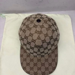 Gucci keps beige - Snygg keps från Gucci i beige och brunt med klassiskt GG-monogram över hela kepsen. Kepsen har en böjd skärm, justerbar rem bak i brunt och en ikonisk röd/grön rand på sidan. Tillverkad i bomullscanvas med läderdetaljer. Perfekt för dig som vill sticka ut med lyxig streetstyle. Helt ny!