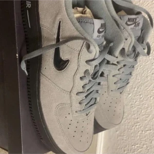 Nike Air Force 1 i grå mocka - Nike Air Force 1 sneakers i ljusgrå mocka med svart sula och svart glansig swoosh. Klassisk siluett med perforerad tå, snörning och låg profil. Perfekta för dig som gillar stilrena och tidlösa sneakers med en twist.