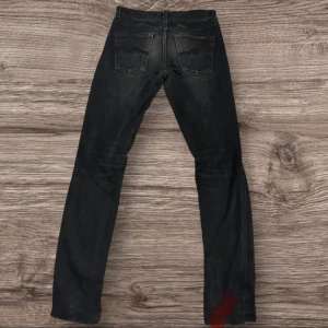 Svarta jeans från Nudie Jeans Co W28 L32 - Svarta jeans från Nudie Jeans Co i klassisk femficksmodell. Jeansen har raka ben och normal passform, med subtila slitningar för en cool look. Tillverkade i jeansmaterial och passar perfekt till en avslappnad stil. Nypris 1599kr