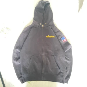 Svart hoodie Indian Fruit of the Loom - Svart hoodie med dragkedja från Fruit of the Loom. Broderad gul Indian-logga på bröstet och stor logga med vinge på ryggen. Amerikansk flagga på ena ärmen. Klassisk känguruficka och huva med metallöljetter.