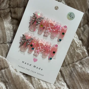 Rosa glittriga lösnaglar med fjärilar - Säljer ett set rosa lösnaglar med glittrig finish, dekorerade med pärlor, färgglada stenar, vita linjer och stora silvriga fjärilar. Naglarna har även små rosetter och hjärtformade detaljer för en extra söt look.