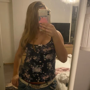 Mörkblommig linnetopp med rosett - Snygg mörkblå linnetopp med blommigt mönster i rosa och lila toner. Toppen har smala axelband och en liten söt rosett framtill vid urringningen. Perfekt för varma dagar och enkel att matcha med jeans eller kjol.