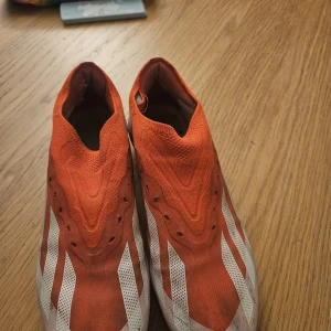 Adidas orange fotbollsskor utan snörning - Säljer ett par orangea och vita fotbollsskor från Adidas med klassiska tre ränder på sidorna. Skorna har en strumpliknande passform utan snörning och mönstrad sula med fasta dobbar för bra grepp på planen. Ovandelen är i syntetmaterial och mesh för lätt känsla.