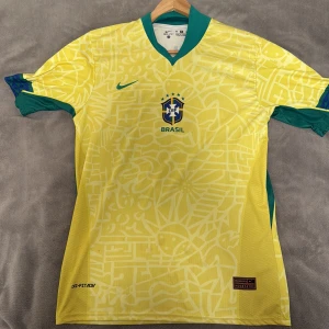 Brasilien Vinicius Jr matchtröja Nike M - Gul Brasilien fotbollströja från Nike med gröna detaljer, officiellt CBF-märke och nummer 20 Vinicius Jr på ryggen. Tillverkad i Dri-FIT-material som andas, har korta ärmar och unikt grafiskt mönster över hela tröjan. Perfekt för dig som älskar landslagsfotboll.