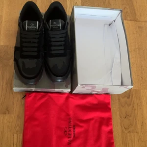 Valentino Garavani Rockrunner sneakers - Snygga svarta Valentino Garavani Rockrunner sneakers med coola camouflagedetaljer. Skorna har en mix av skinn och textil, klassisk snörning och ikoniska nitar på sulan. Kommer med originalkartong och röd dustbag. Perfekta för dig som vill ha en exklusiv och stilren look.