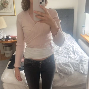 Ljusrosa omlottopp från Stradivarius - Supersöt ljusrosa omlottopp från Stradivarius i ribbad bomullsmix. Toppen har långa ärmar, croppad passform och knytband i midjan. Perfekt för dig som gillar en feminin och trendig look med v-ringning och fina volangdetaljer vid ärmslut och nederkant.