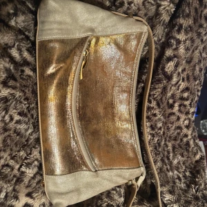 Guldig axelväska i skinnlook från Coach London - Snygg axelväska i guldig metallic med beige detaljer och axelrem. Väskan har flera fack med dragkedja och insida i ljus textil. Perfekt för dig som vill sticka ut med en glansig accessoar. Guldiga metalldetaljer och stilren design.