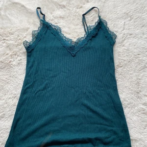 Mörkgrön spetslinnetopp från Amisu - Supersöt mörkgrön linnetopp från Amisu med tunna axelband och vacker spetsdetalj vid bysten. Toppen är ribbad och har en lätt vågig kant nertill. Perfekt för dig som gillar en feminin och trendig look.