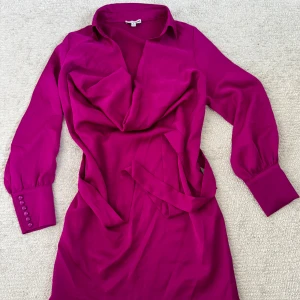 Cerise skjortklänning från Fashion Nova - Säljer en cerise skjortklänning från Fashion Nova med långa ärmar, breda manschetter och knappar vid ärmslut. Klänningen har en snygg omlott-effekt framtill, krage och tillhörande bälte i midjan. Perfekt för dig som vill sticka ut med färg och stil.