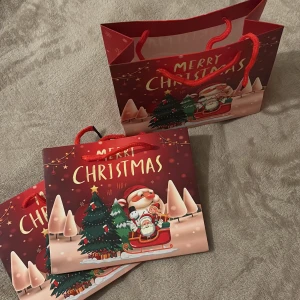 Julig shoppingväska med tomtemotiv - Säljer en röd shoppingpåsar med julmotiv, där tomten och en gran syns tillsammans med texten 'Merry Christmas'. Påsar har röda snören som handtag och är perfekt för att ge bort presenter i under julen.