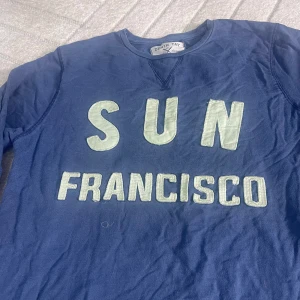 Blå långärmad tröja Denim Day M - Blå långärmad tröja från Denim Day i storlek M med stora vita bokstäver 'SUN FRANCISCO' framtill. Tröjan har rund halsringning och är tillverkad i mjuk bomull. Perfekt för en avslappnad och cool stil.
