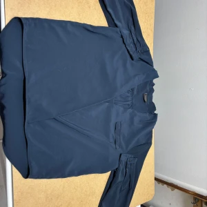 Mörkblå blus från Vila med v-ringning - Snygg mörkblå blus från Vila med v-ringad hals och långa ärmar. Blusen har en enkel, stilren design med knappdetaljer vid ärmsluten och ett litet bröstfack. Perfekt för dig som gillar en clean och modern look.