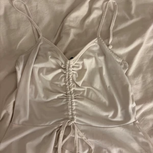 Vit omlottklänning med snörning - Supersöt vit linne med tunna axelband och snygg snörning framtill. Linnet har omlott-detalj och volang nedtill som ger en extra feminin touch. Materialet är mjukt och har en lätt glansig finish, perfekt för varma dagar.