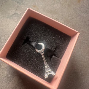 Silverfärgat berlock med Eiffeltorn - Snyggt berlock  med ett silverfärgat Eiffeltorn som hänge. Hängsmycket har fina detaljer och sitter på ett svart band. Perfekt för dig som gillar Paris eller vill ha något unikt och stilrent.