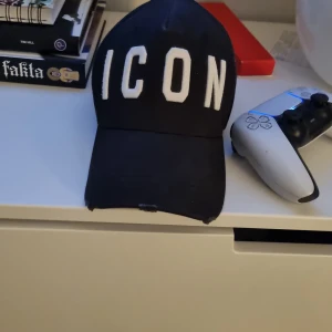 Svart ICON keps från Dsquared2 - Snygg svart keps från Dsquared2 med stor vit broderad text 'ICON' framtill. Klassisk böjd skärm och justerbar passform. Perfekt för dig som vill sticka ut med en trendig accessoar. Materialet är bomull och kepsen har en cool streetstil.