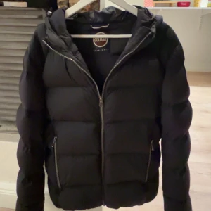 Colmar jacka - Säljer en svart pufferjacka från Colmar med huva och två dragkedjefickor. Jackan har en clean look med Colmar-logga på ärmen och grova kanaler. Perfekt för kalla dagar och riktigt snygg till streetwear-stilen.