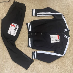 Svarta mjukisbyxor DSQUARED2 x Ibrahimovic - Svarta mjukisbyxor från DSQUARED2 x Ibrahimovic ICON-kollektionen. Byxorna har vit och röd grafisk print på ena benet, elastisk midja med dragsko och dragkedja vid benslut. Snygg sportig look med bekväm passform och coolt tryck.