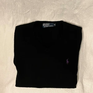 Svart v-ringad tröja Polo Ralph Lauren - Svart v-ringad tröja från Polo Ralph Lauren i 100% merinoull. Klassisk design med lila broderad logga på bröstet. 
